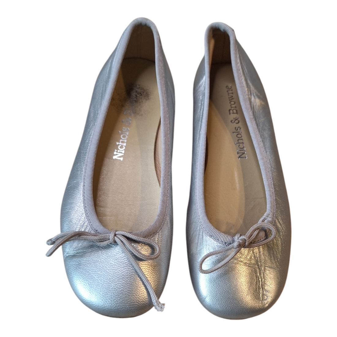 A pair of Nichols &amp; Browne Silver Leather Ballerinas, size 30. The ballerinas are made of shiny silver leather with a light gray trim and a small bow at the toe. The interior lining is light tan.